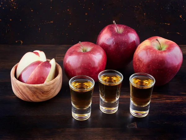 Friss piros almák és alma pálinka poharak egy sötét fafelületen.