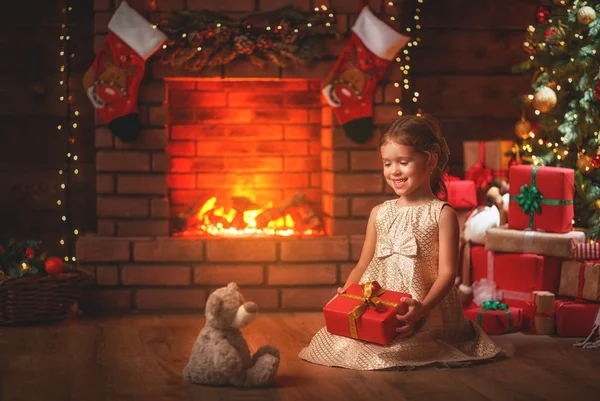 Egy boldog kislány karácsonyi ajándékot tart a kezében, miközben a kandalló előtt ül.